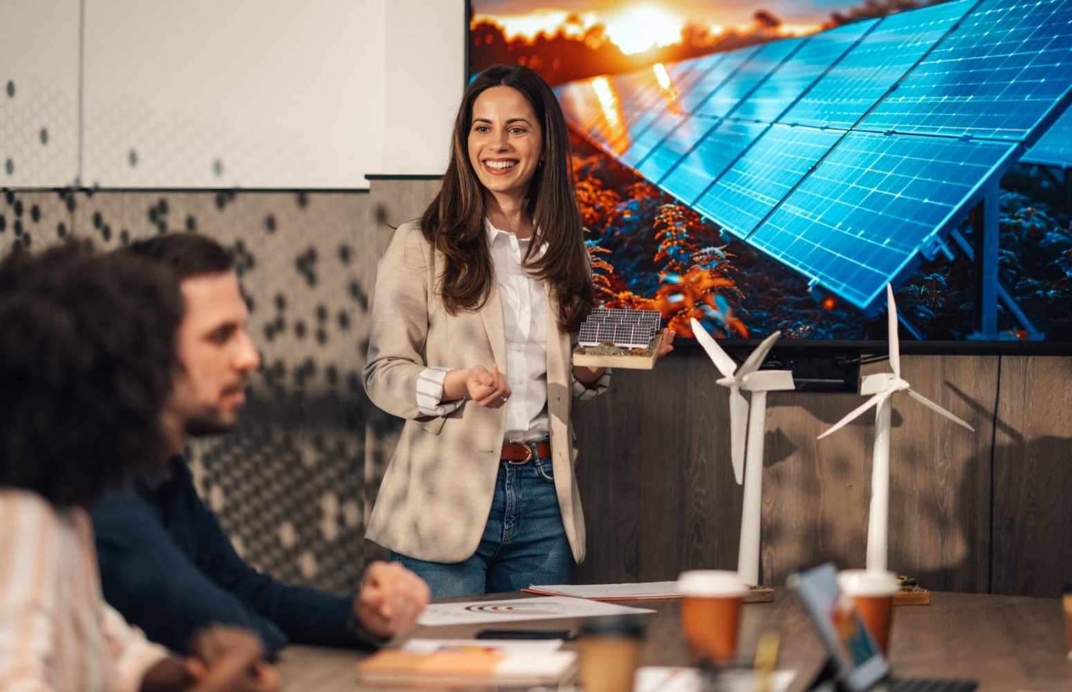 Portrait of smiling businesswoman standing at boardroom with solar panel model in hands and having presentation in front of diverse team of experts. Ecologist presenting innovative technology to staff